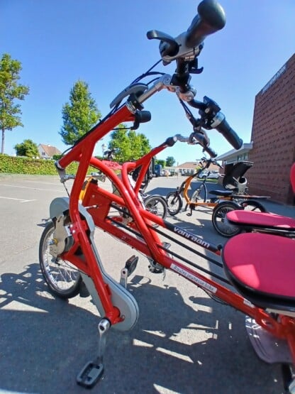 Vélo adapté Van Raam, rouge, stationné, avec sièges rouges, dans un cadre extérieur ensoleillé.
