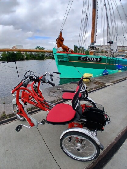 Vélo adapté Van Raam rouge à deux places au bord de l'eau, avec un bateau en arrière-plan.