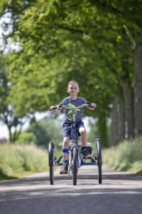 Un garçon souriant pédale sur un tricycle bleu sur une route bordée d'arbres.