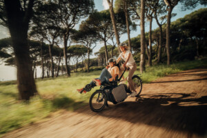 Une femme roule sur un vélo adapté avec un enfant à l'arrière, entourés de nature.