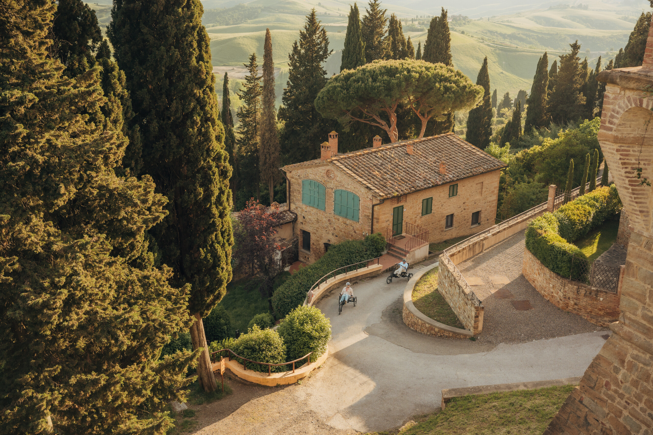 Deux personnes sur des vélos adaptés, roulant sur un chemin entouré de verdure près d'une maison en pierre.