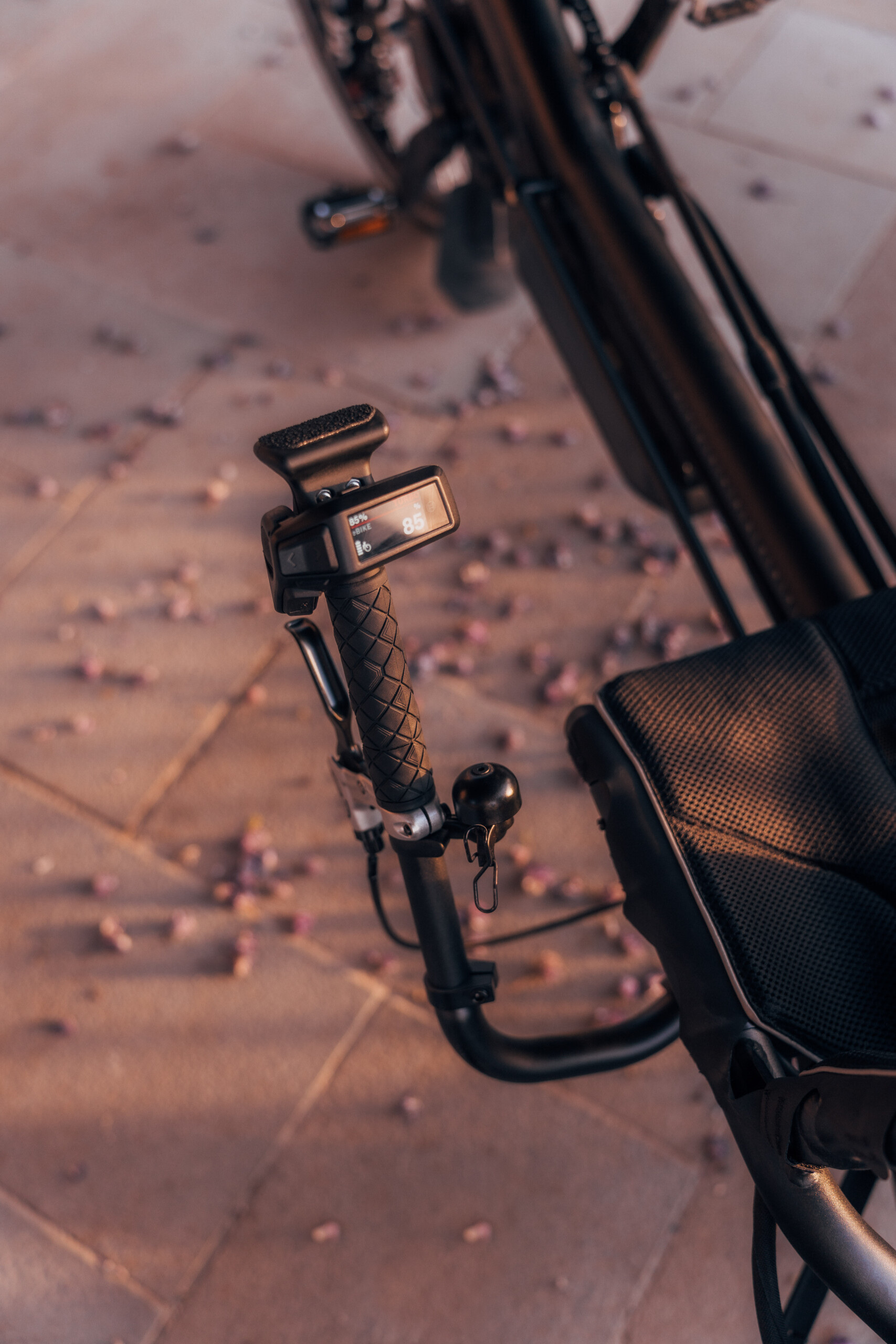 Détail d'un contrôleur de vélo électrique avec écran, guidon et sonnette, sur un sol orné de pétales de fleurs.