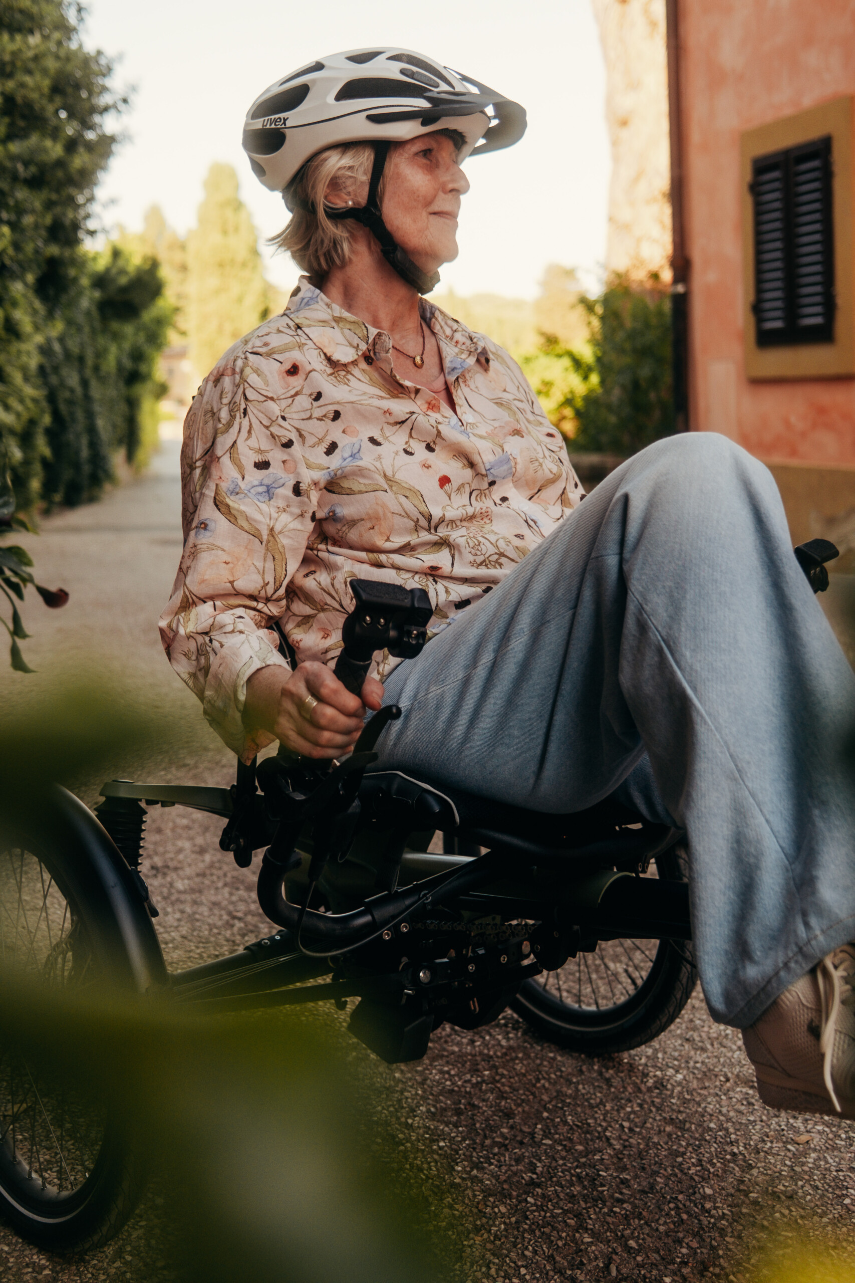 Femme âgée sur un vélo adapté, portant un casque, dans un chemin bordé de verdure.