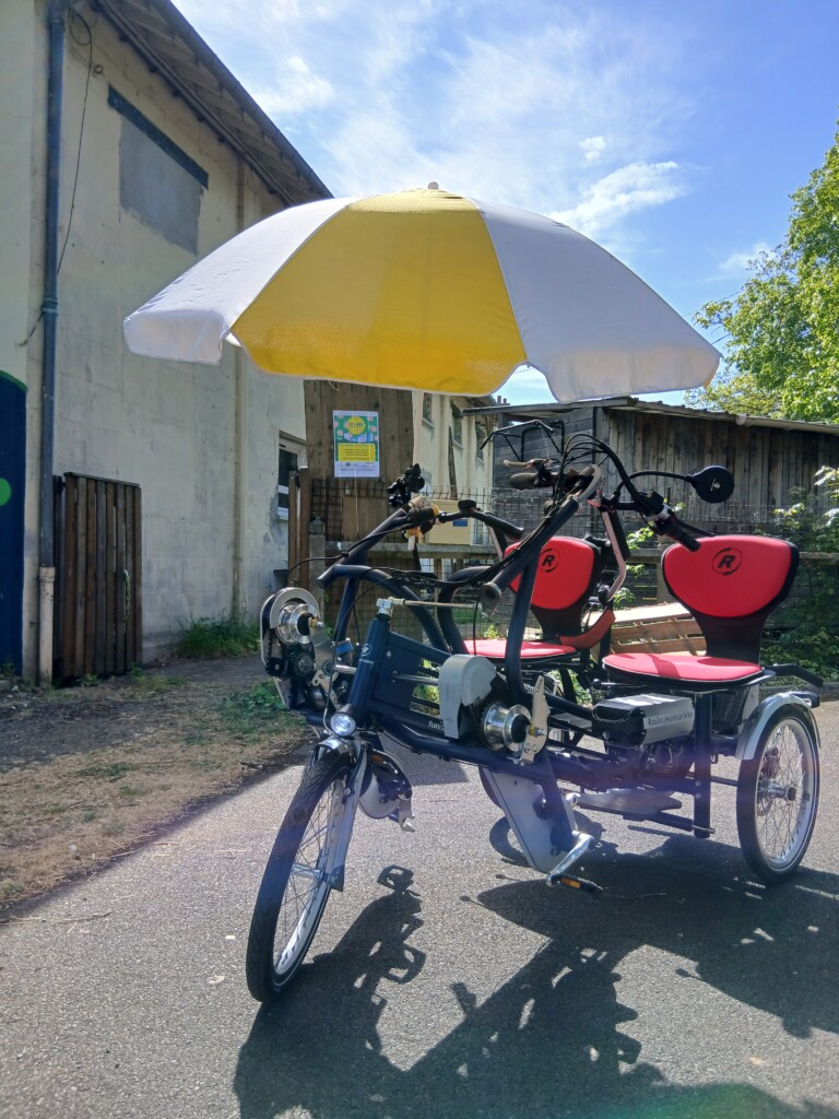Vélo adapté avec deux sièges, parasol jaune, sur une route, proposé par Roulez JEUnesse Loisirs.