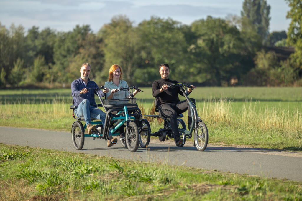 Trois personnes souriantes circulant sur des vélos adaptés dans un paysage verdoyant.