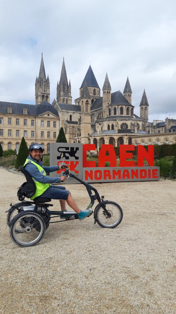 Homme souriant sur un vélo adapté devant le panneau "CAEN NORMANDIE" et un bâtiment historique.