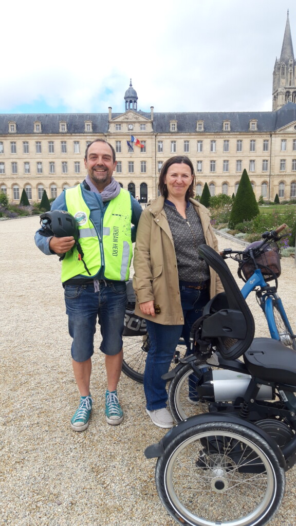 Deux personnes souriantes posent avec des vélos près d'un bâtiment historique, promotionnant des solutions de mobilité durable.