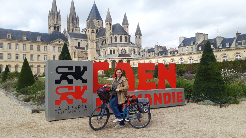 Femme posant sur un vélo devant le panneau "CAEN NORMANDIE" avec un bâtiment historique en arrière-plan.