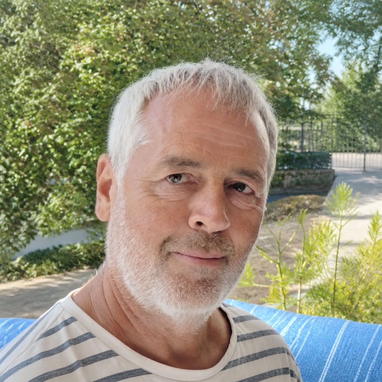 Homme souriant en extérieur, avec des cheveux gris, près d'une végétation verdoyante, sur un fond de fauteuil bleu.